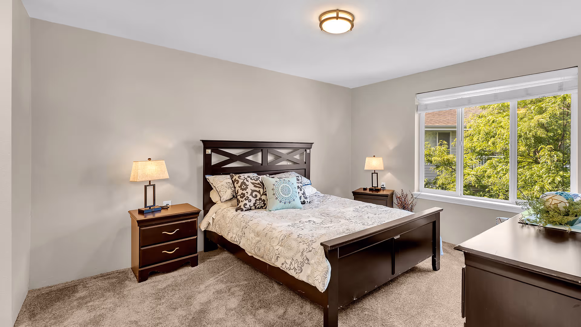A bedroom with a large window showing green trees outside. The room features a dark wooden bed frame with a patterned bedspread and multiple decorative pillows. There are two matching dark wooden nightstands on either side of the bed, each with a lamp. A dark wooden dresser is visible on the right side of the image. The walls are painted light gray and the floor is carpeted in a neutral tone.