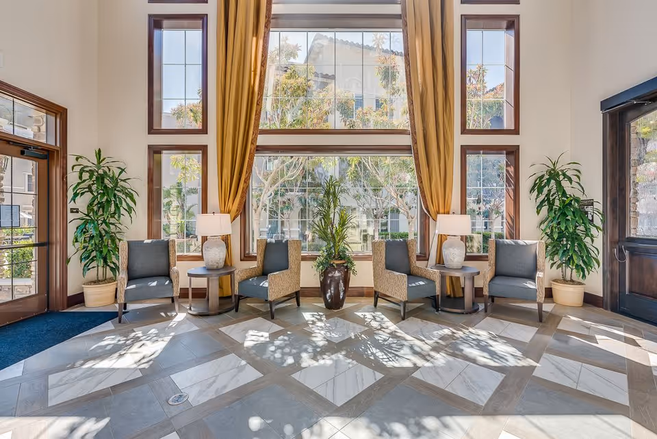 A bright and spacious seating area with four cushioned armchairs arranged in pairs around two small round tables with lamps. Large windows with wooden frames and golden curtains allow natural light to fill the room. Two large potted plants flank the seating area, and a tall plant in a decorative pot is centered between the chairs. The floor features a patterned tile design.