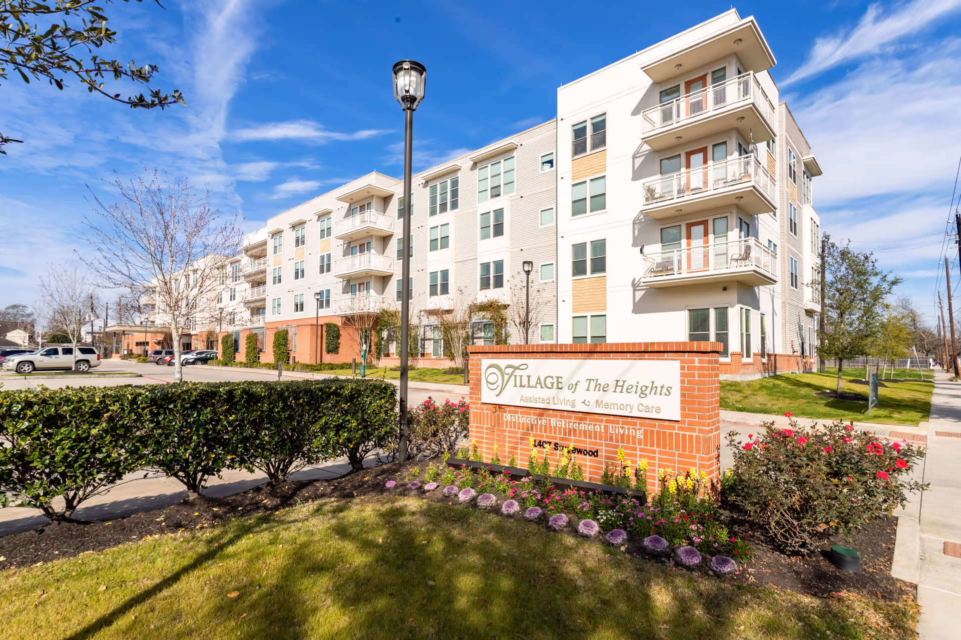Exterior view of a multi-story assisted living and memory care facility named Village of the Heights. The building is modern with balconies and large windows. In front, there is a brick sign with the facility's name and landscaping with bushes and flowers. A parking lot and street are visible to the left, and a sidewalk runs along the right side of the building under a blue sky with some clouds.