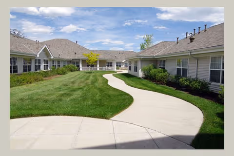 Curved concrete walkway surrounded by well-maintained green grass and shrubs, leading between single-story buildings with beige siding and multiple windows under a partly cloudy sky.