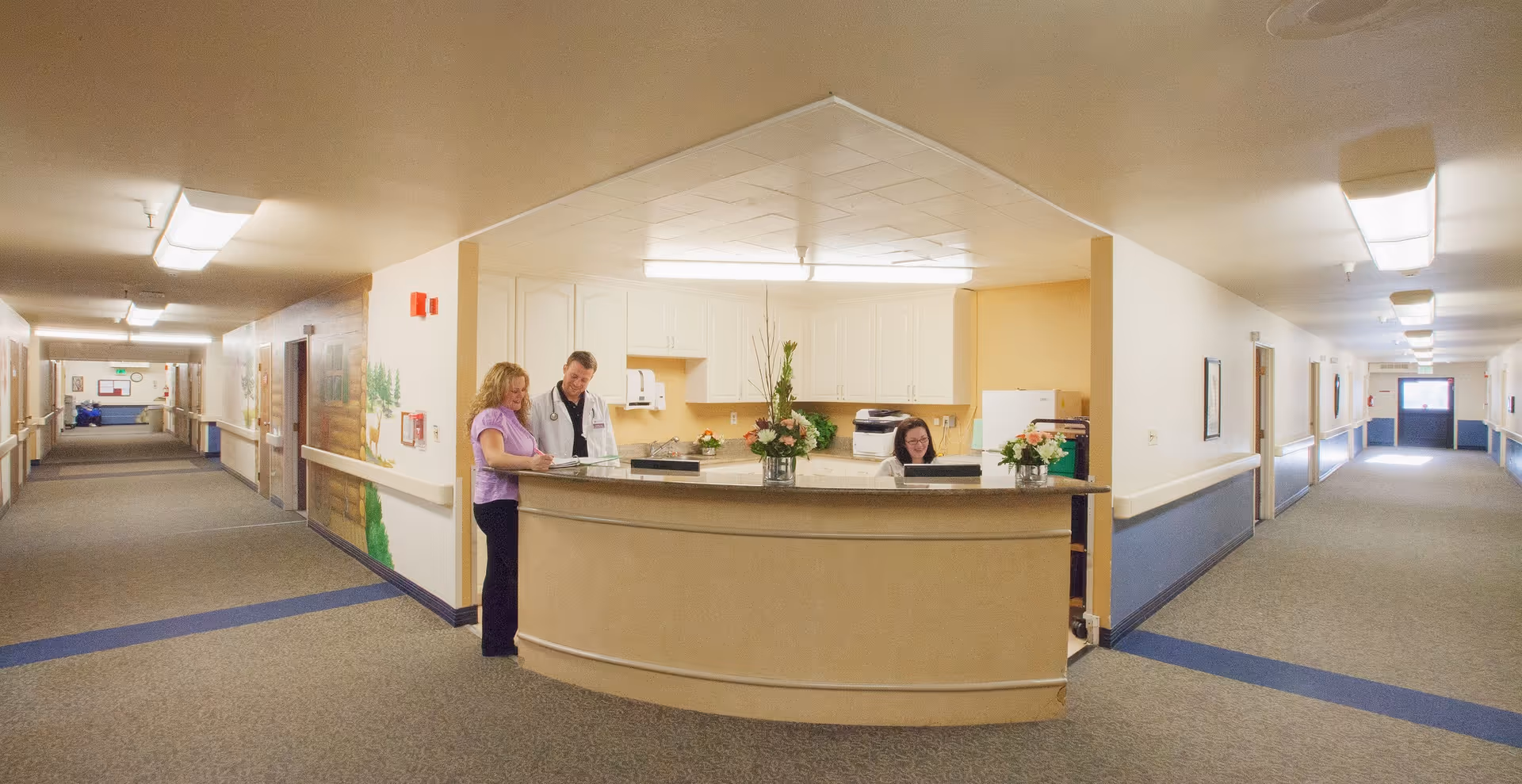 Reception desk area in a senior living facility hallway with two staff members standing and one sitting behind the desk. The hallway extends on both sides with doors and handrails along the walls.