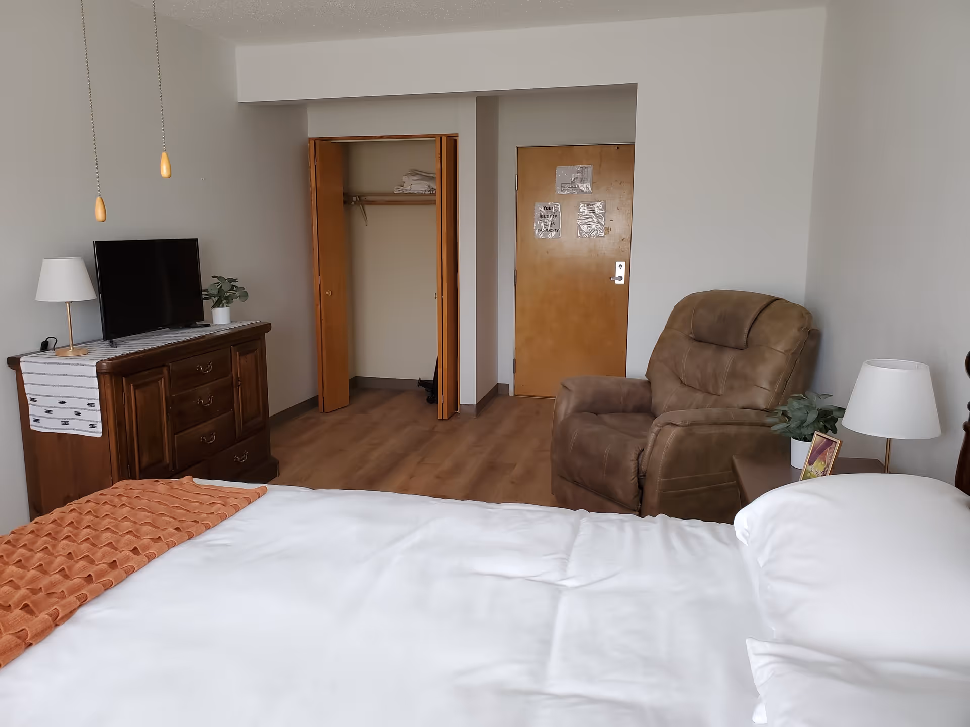A bedroom in a senior living facility featuring a bed with white linens and an orange blanket, a wooden dresser with a TV and lamp on top, an open closet with shelves and hangers, a brown recliner chair, a side table with a lamp and a small plant, and a wooden door with notices attached.