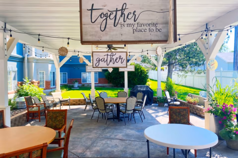Outdoor covered patio area with multiple tables and chairs arranged for seating. Decorative signs hang from the ceiling with phrases like 'together is my favorite place to be' and 'gather'. String lights and hanging decorations add ambiance. Surrounding the patio are plants, greenery, and a building with windows.