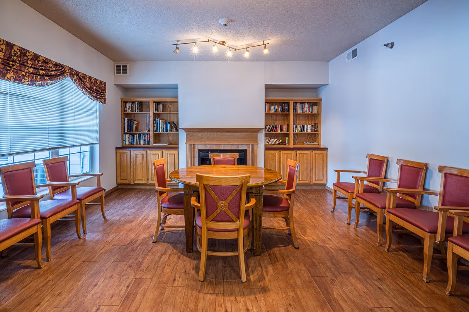 A senior living common room with a round wooden table surrounded by chairs, bookshelves, and a fireplace.