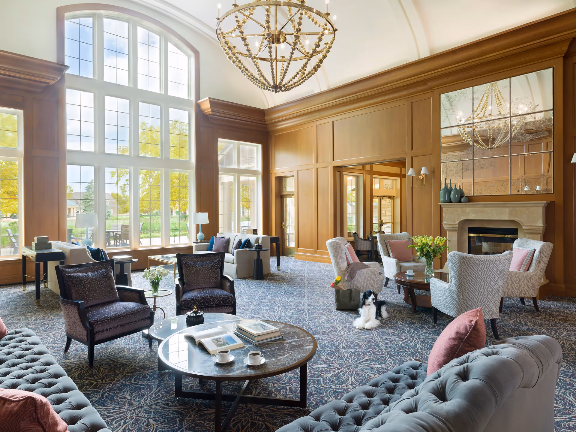 A spacious and elegant living room with large arched windows letting in natural light. The room features comfortable seating including armchairs and sofas with pink and purple cushions, a round coffee table with books and cups, a fireplace with a large mirror above it, and a decorative chandelier hanging from the ceiling. A small black and white dog is lying on the carpet near the chairs.