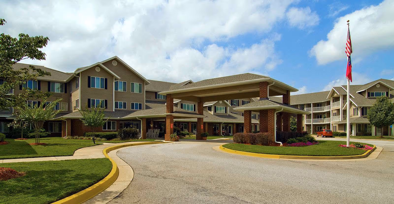 Exterior front entrance of Holiday Pinegate senior living facility with a covered porte-cochere, circular driveway, and flagpoles.