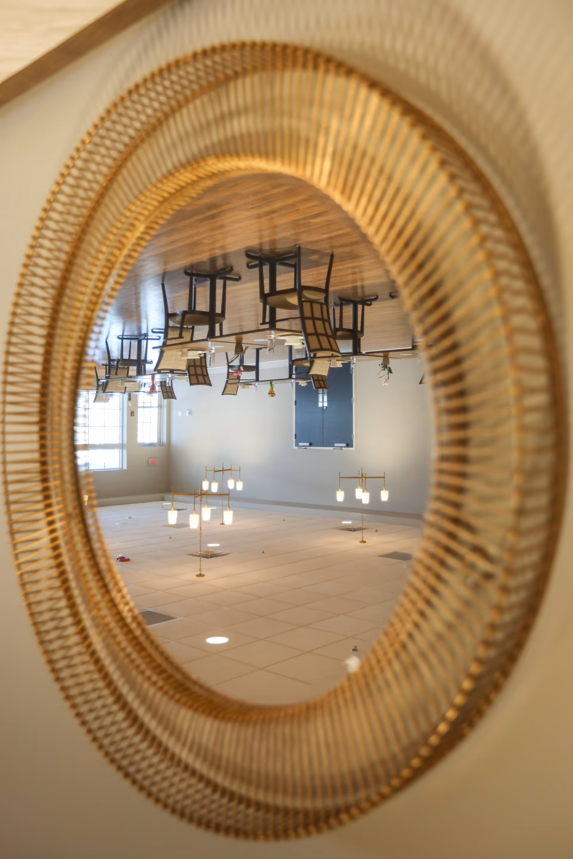 Reflection in a round, gold-framed mirror showing an empty dining area with wooden floor, tables, and chairs. The ceiling with lights and vents is also visible in the reflection.