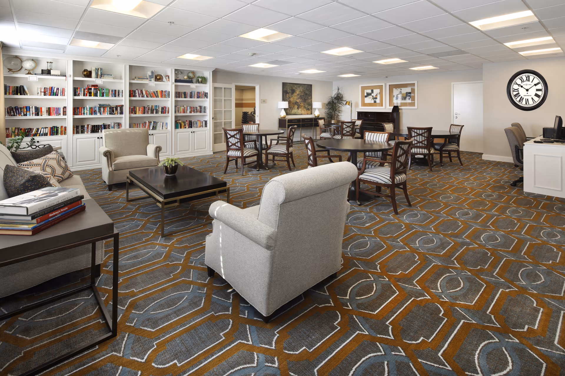 A spacious and well-lit common area in a senior living facility featuring comfortable armchairs, a sofa, round tables with chairs, a large bookshelf filled with books, decorative items, a piano, and a wall clock. The room has a patterned carpet and neutral-colored walls with framed artwork.