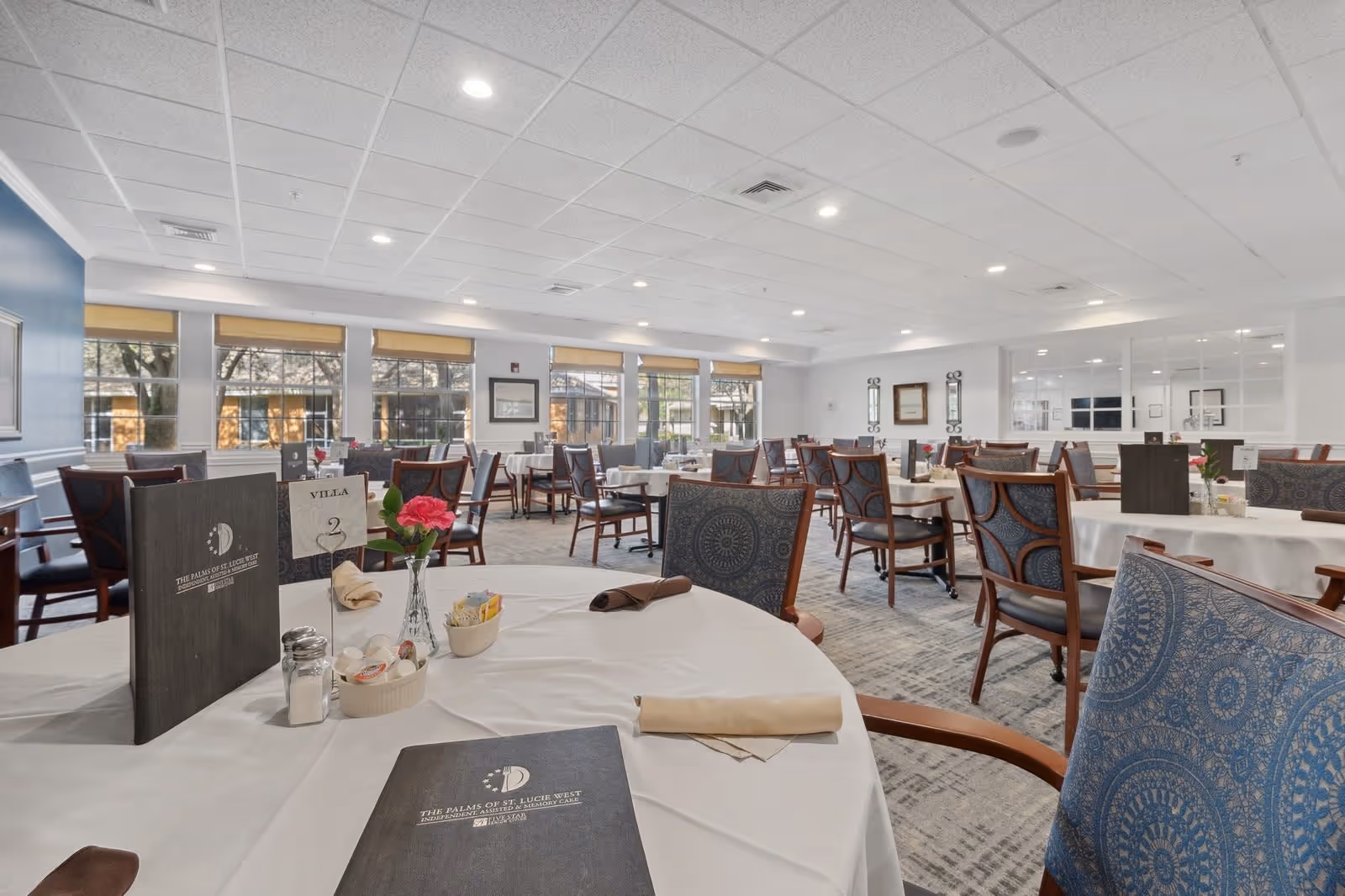A spacious dining room with multiple round tables covered in white tablecloths, each set with menus, napkins, and small flower vases. The room has large windows with beige blinds, blue patterned chairs, and a carpeted floor. The atmosphere is bright and inviting with recessed ceiling lights.