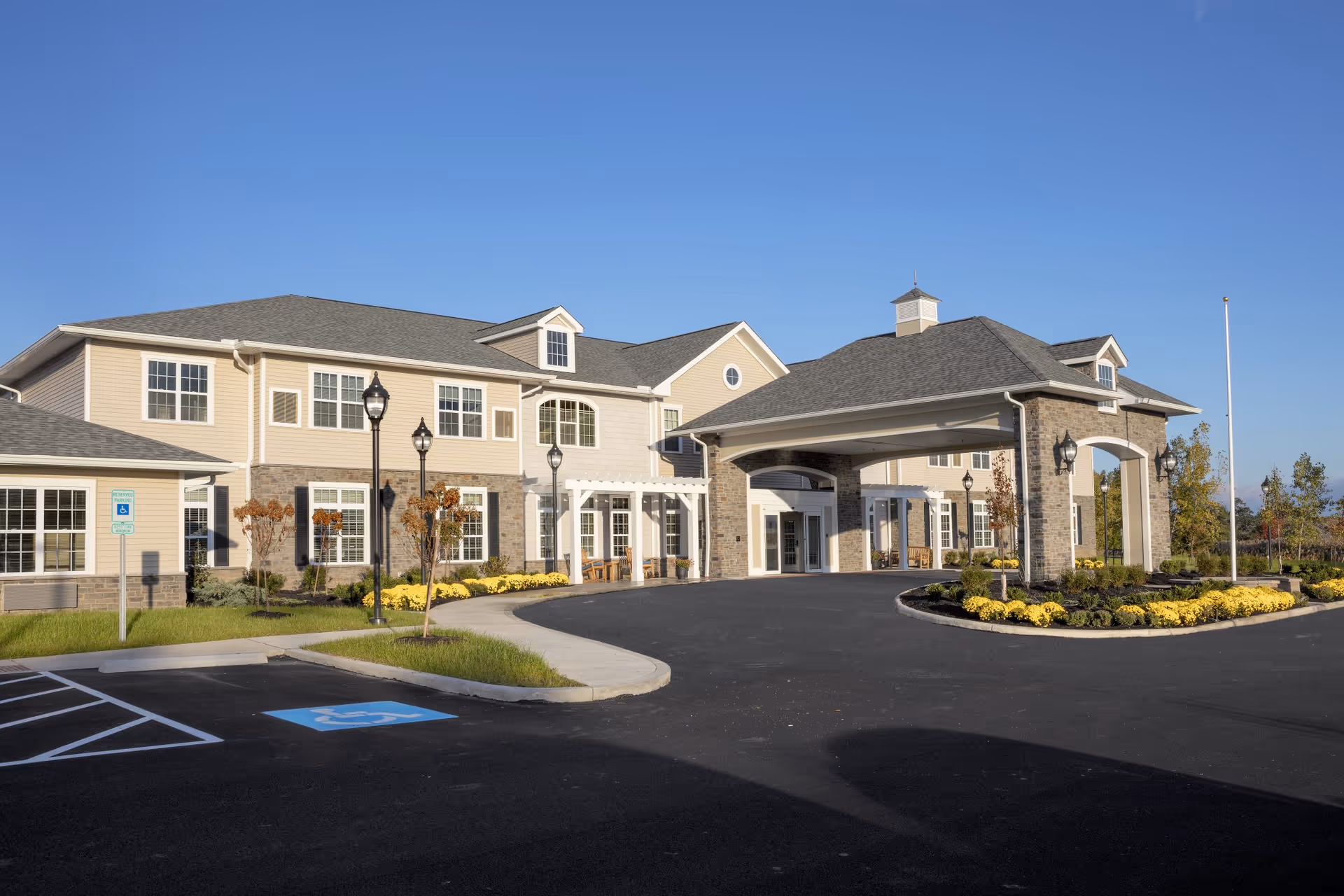 Exterior view of Brooklyn Pointe Assisted Living & Memory Care building with a covered entrance, multiple windows, landscaped flower beds with yellow flowers, and a clear blue sky.