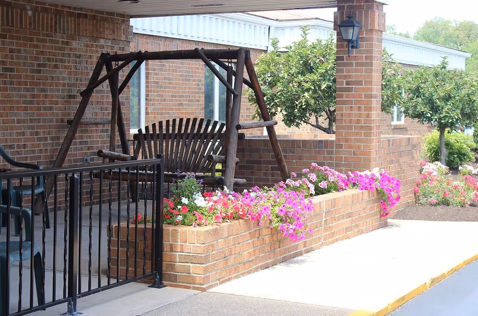 A covered outdoor patio area with a wooden swing bench, surrounded by brick walls and flower beds filled with colorful flowers. There are also some green plastic chairs and a black metal railing. The background shows a brick building and some small trees.