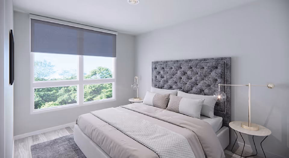 A modern bedroom with a large bed featuring a tufted gray headboard, light gray bedding, and multiple pillows. There are two small round bedside tables with lamps on each side of the bed. A large window with a gray roller shade lets in natural light and offers a view of green trees outside. The walls are painted light gray and the floor has a wood-like finish with a gray rug partially visible.