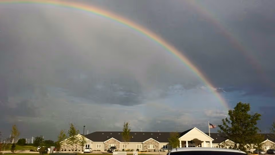 A double rainbow arches over the front of a single-story senior living building with an entrance, flagpole, and parked cars.