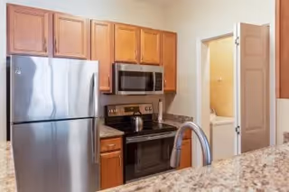 Modern kitchen area with stainless steel refrigerator, microwave, and electric stove. Wooden cabinets are mounted above the countertop, and a granite countertop with a curved faucet is visible in the foreground. A doorway leads to another room with a glimpse of a bathroom.