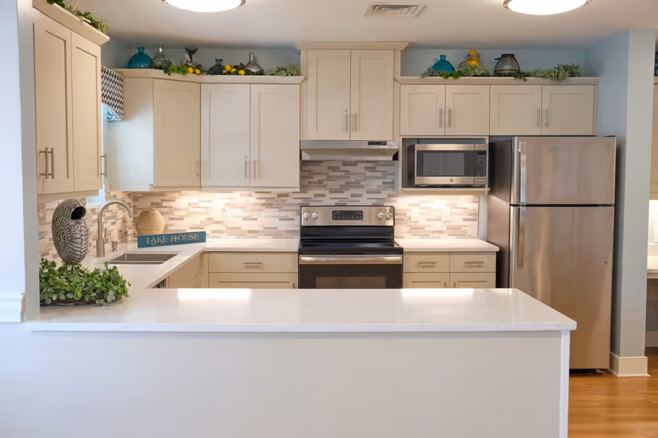A modern kitchen with light-colored cabinets, a stainless steel refrigerator, microwave, and oven. The backsplash features a mosaic tile design in neutral tones. There is a white countertop island in the foreground and decorative greenery and vases on top of the cabinets.