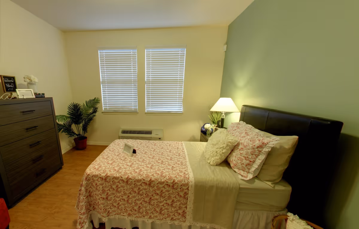 A cozy bedroom with a floral-covered bed, nightstand and lamp, dresser, potted plant, and two windows with blinds.