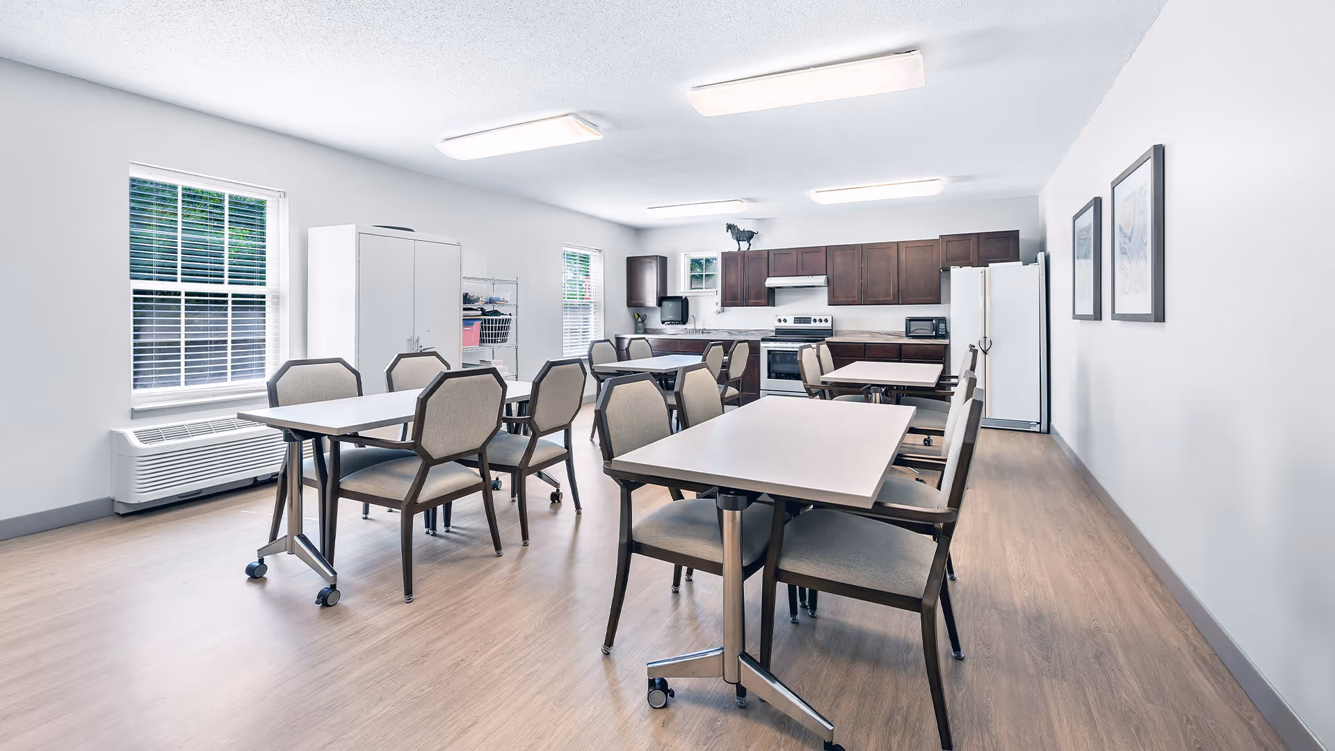 A bright, clean dining area with several tables and chairs arranged neatly. The room features light-colored walls, wood flooring, and two windows letting in natural light. At the far end, there is a kitchen area with dark wood cabinets, a stove, microwave, and refrigerator. Two framed pictures hang on the right wall.