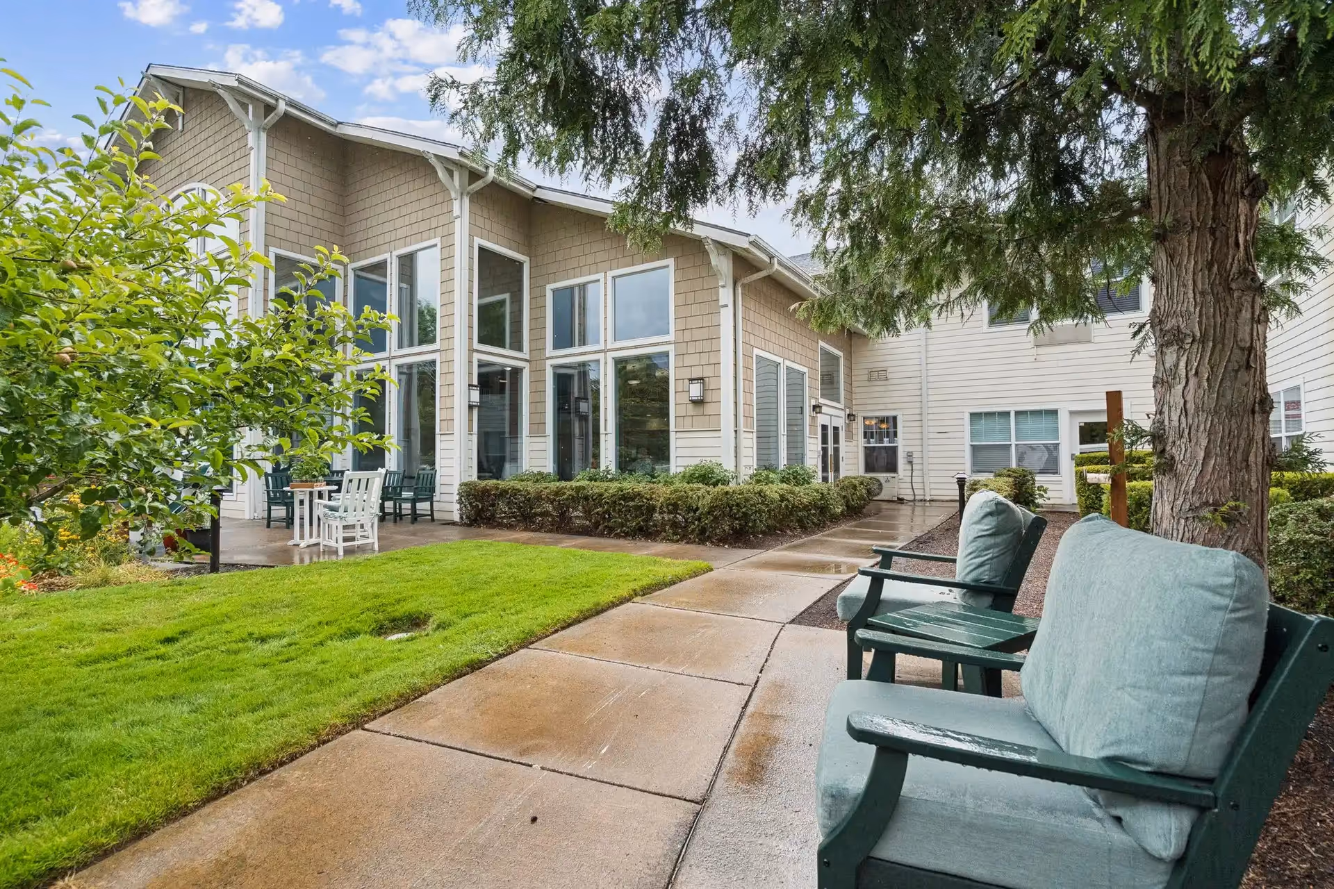 Outdoor seating area at Clearwater Springs by Cogir featuring cushioned green chairs along a paved walkway, surrounded by trees, bushes, and a well-maintained lawn. The building exterior has large windows and beige siding under a partly cloudy sky.