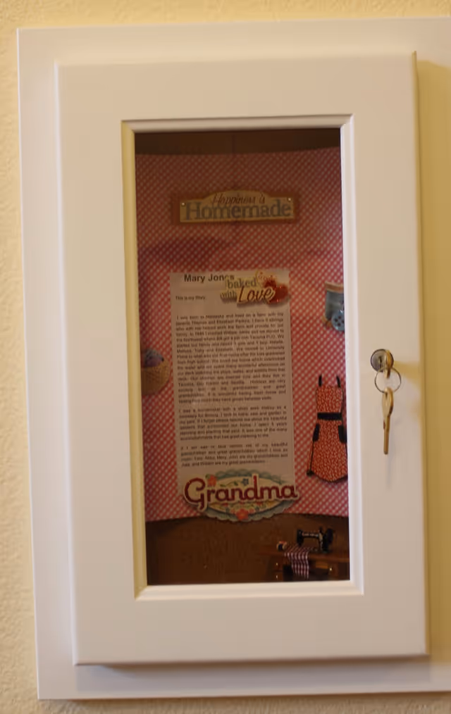 White wall-mounted display cabinet with a glass door showing a pink 'Grandma' homemade-themed poster and a set of keys hanging from the lock.