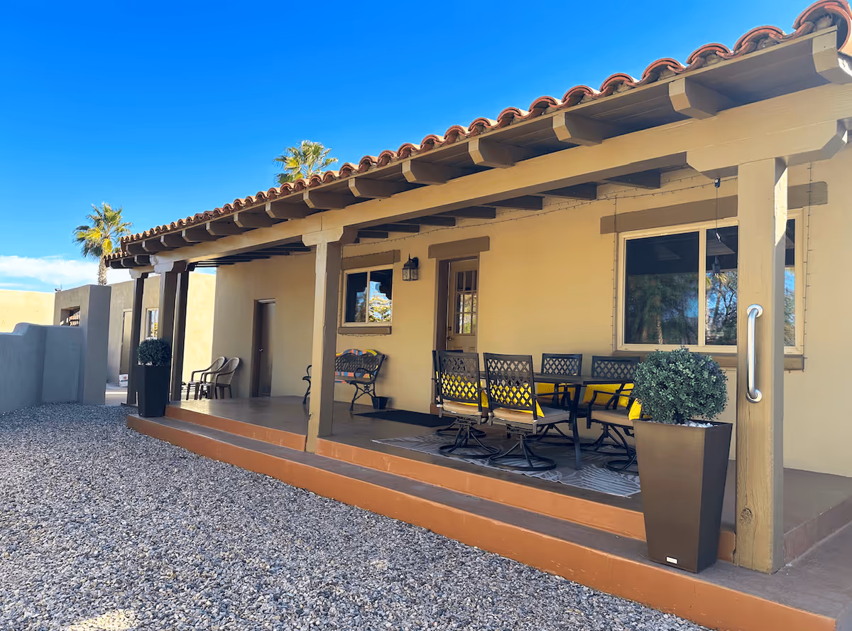 Outdoor covered patio area with a tiled roof and wooden beams, featuring a dining table with six chairs and two potted plants on either side. The ground is covered with gravel, and there are palm trees visible in the background under a clear blue sky.