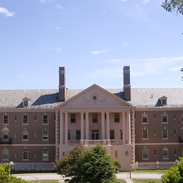Front exterior view of a large, stately brick building with classical architectural features including tall columns and a triangular pediment. The building has multiple windows and two prominent chimneys on the roof. There are trees and greenery in the foreground under a clear blue sky.