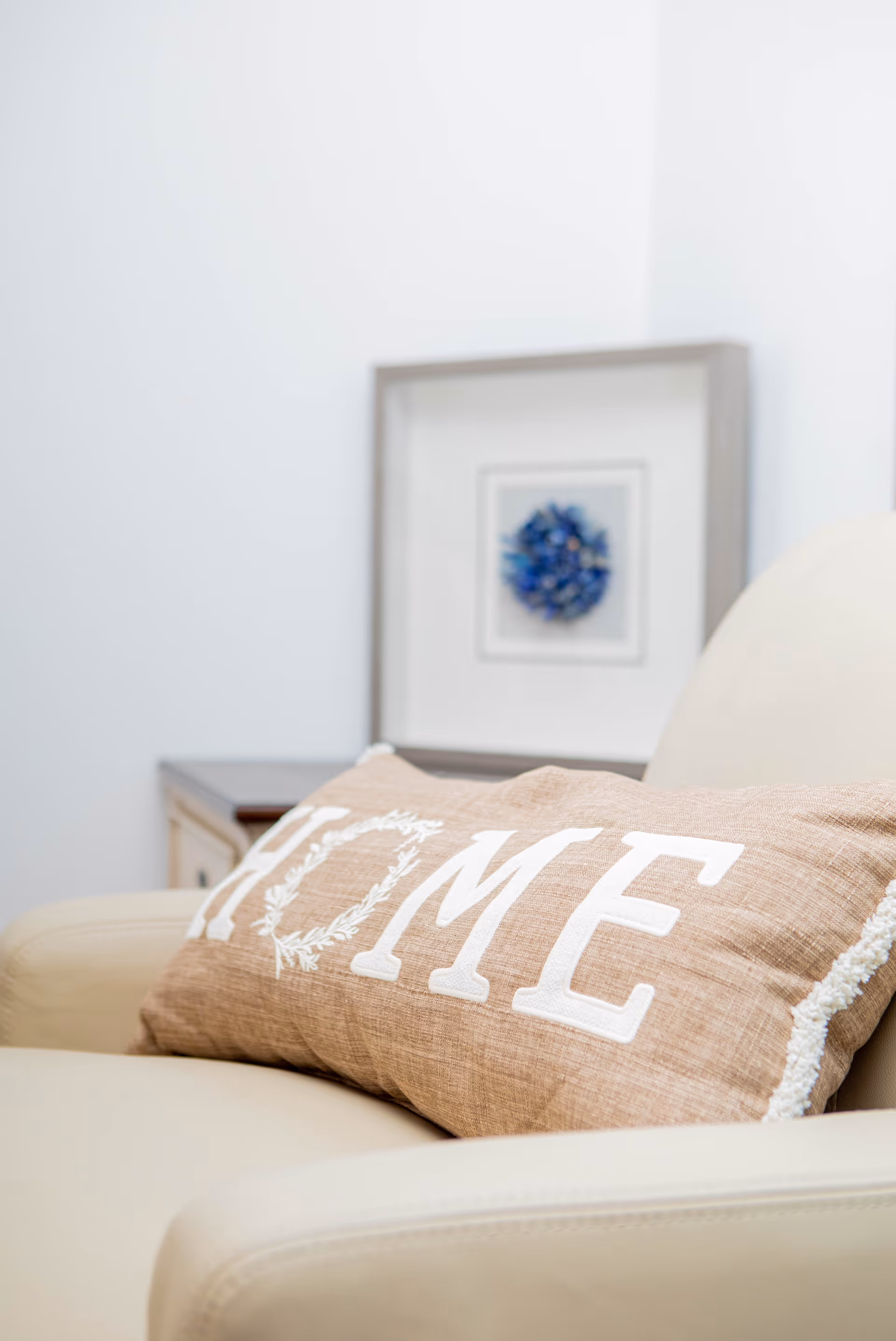 A beige armchair with a brown rectangular pillow that has the word 'HOME' embroidered in white letters. In the background, there is a framed piece of art with blue floral design on a white wall.