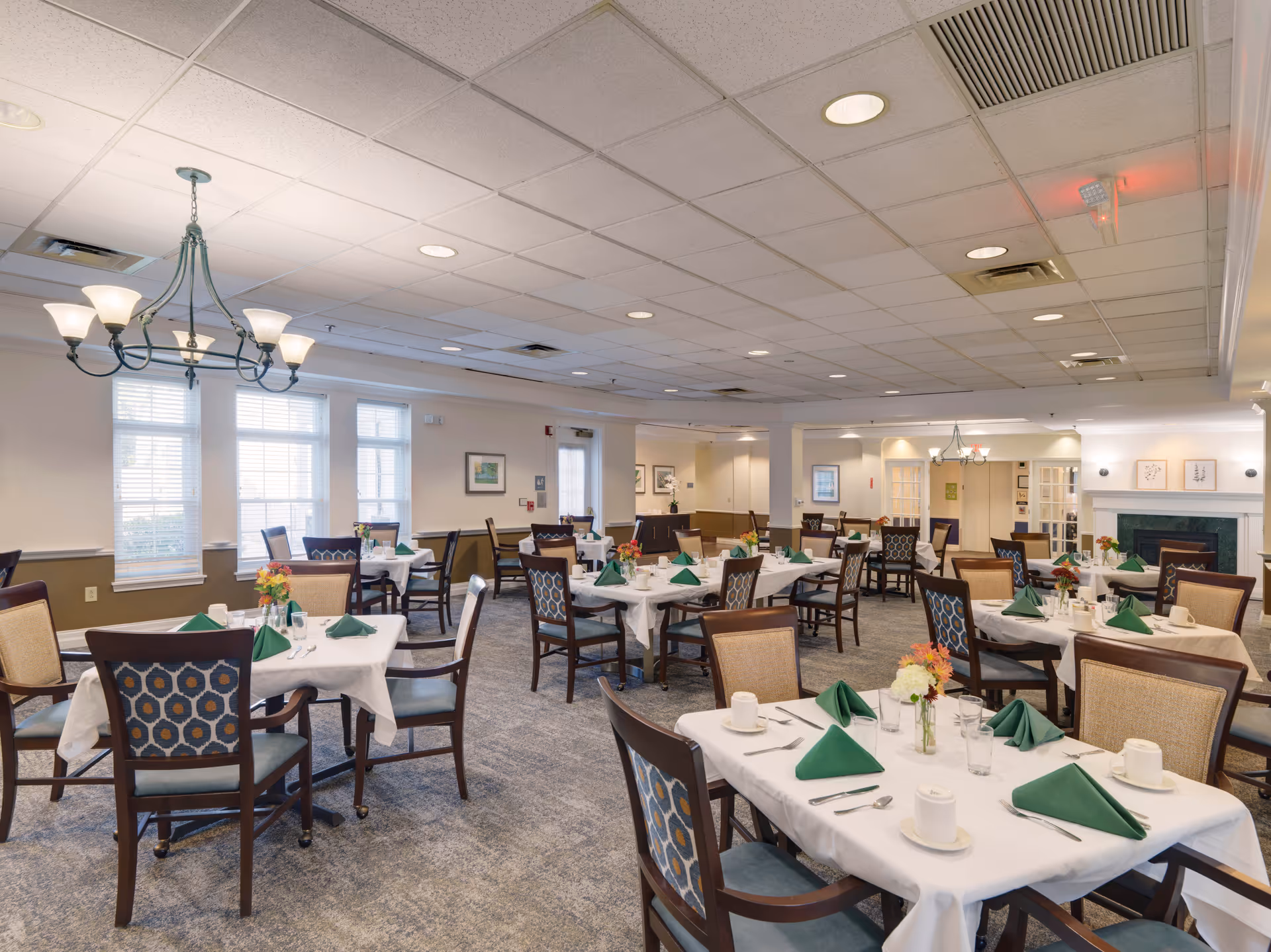 A spacious dining room with multiple tables covered in white tablecloths, each set with green folded napkins, cups, glasses, and silverware. The room features patterned chairs, large windows letting in natural light, a chandelier, and a fireplace in the background.