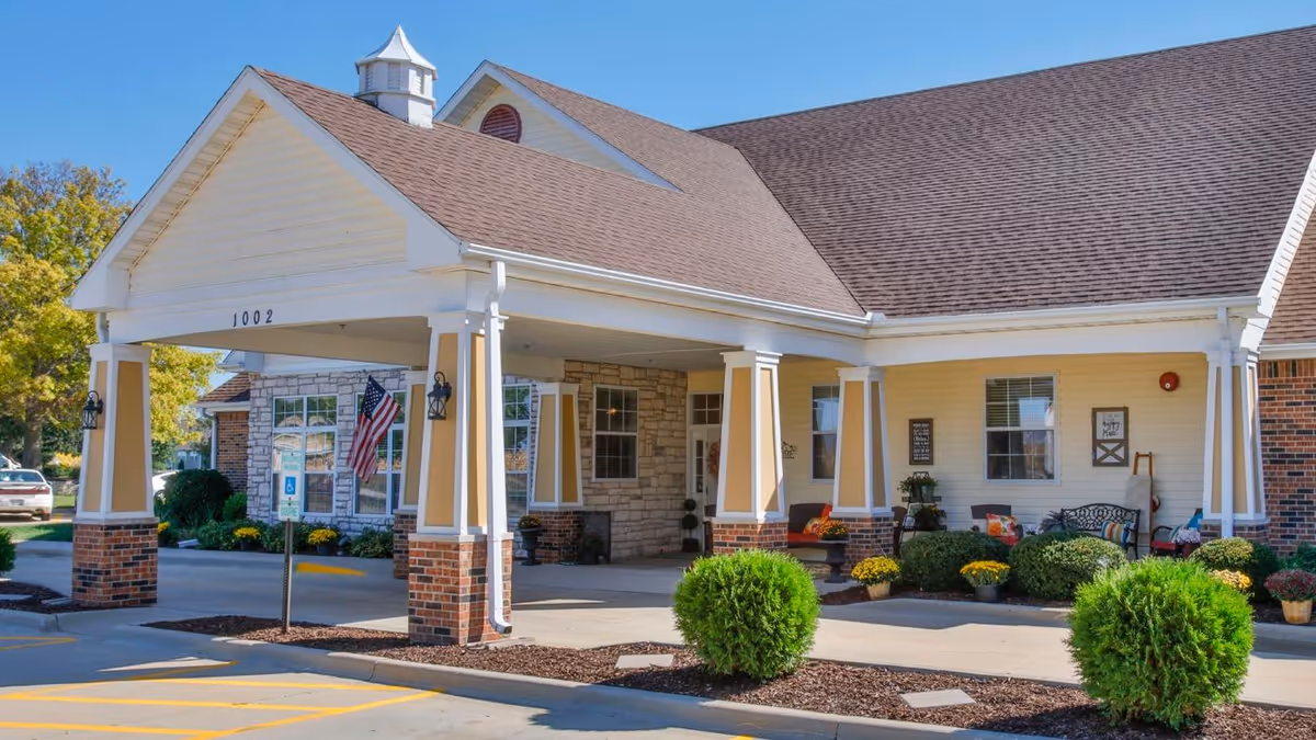 Exterior view of Bickford of Champaign senior living facility showing the entrance with a covered drop-off area supported by columns. The building has a combination of brick and siding with a pitched roof. There are shrubs and potted plants near the entrance and an American flag is displayed near the door.
