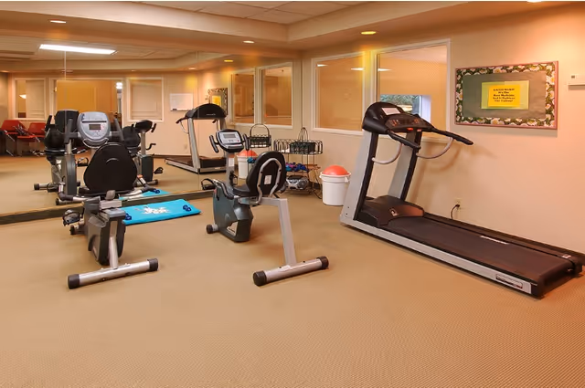 A small fitness room with exercise equipment including a treadmill, two stationary bikes, and a large wall mirror reflecting the room. The walls are beige, and there is a bulletin board with a yellow notice on the right wall. The floor is carpeted, and there is a white trash bin with a red lid near the treadmill.