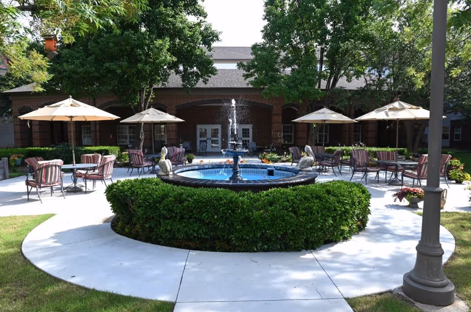 Outdoor courtyard area with a central water fountain surrounded by neatly trimmed bushes. Several tables with umbrellas and cushioned chairs are arranged on a concrete patio. Trees and a brick building with multiple windows and doors are visible in the background.