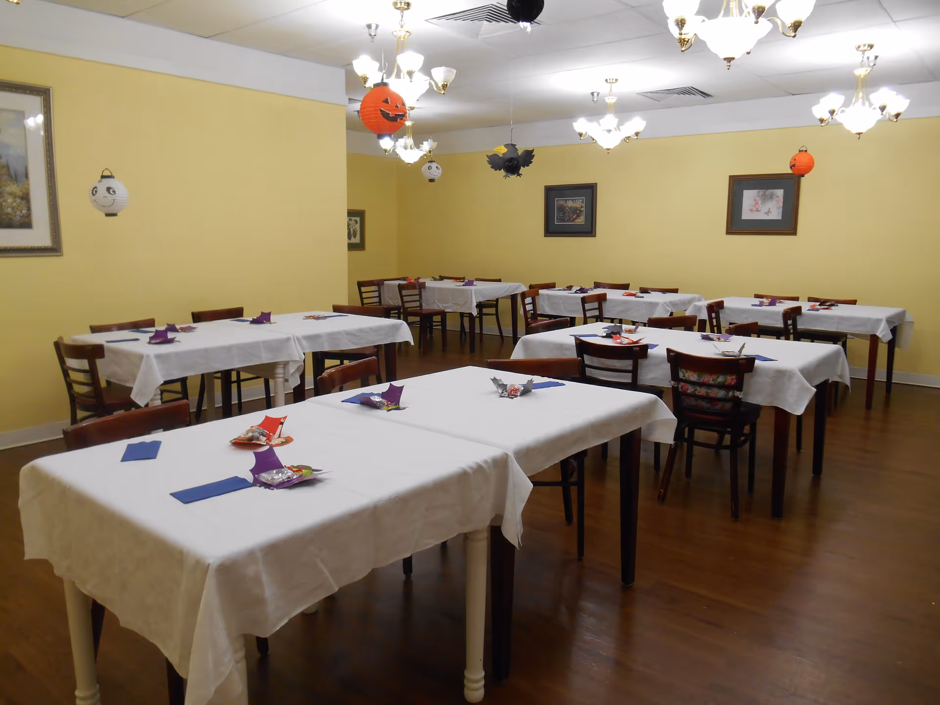 Dining room with several tables draped in white tablecloths, wooden chairs, and hanging Halloween decorations.