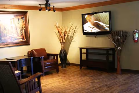 Cozy senior facility living room with seating, a wall-mounted TV, and decorative vases.
