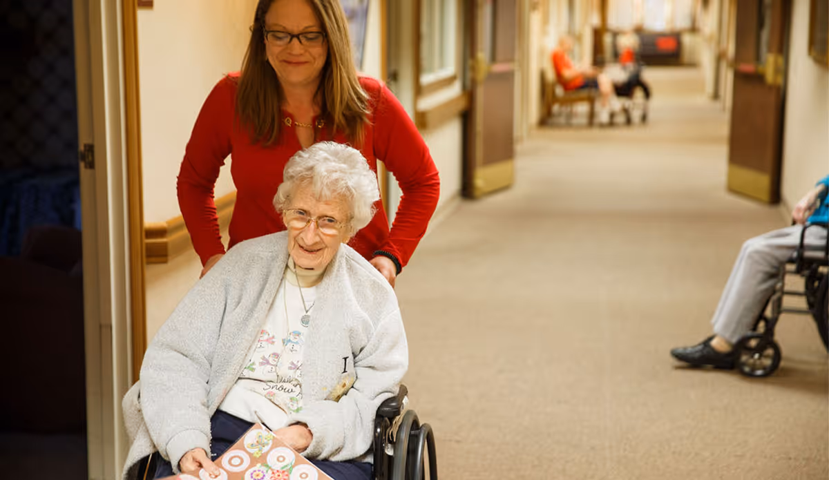 An elderly woman in a wheelchair smiling while being pushed by a caregiver in a red sweater down a hallway of a senior living facility. Other residents in wheelchairs are visible further down the hallway.