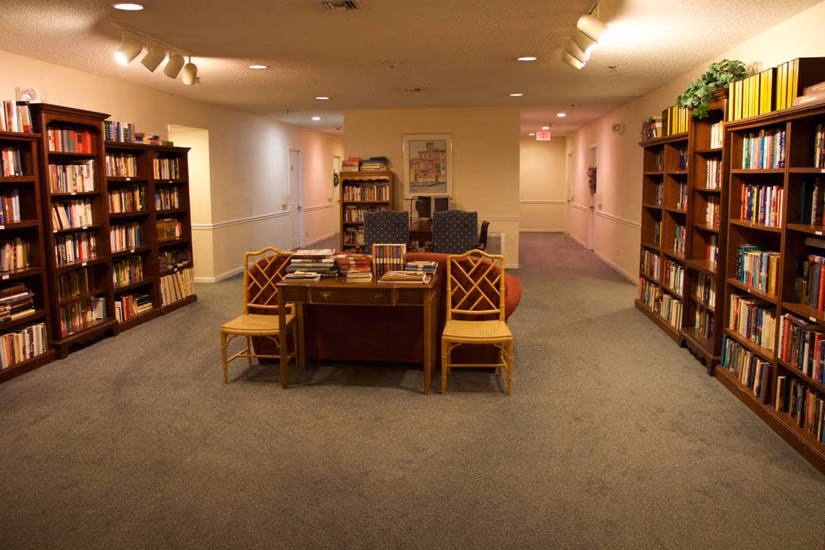 Interior view of a quiet library or reading area in a senior living facility with bookshelves filled with books lining both sides of the room. In the center, there is a wooden table with stacks of books and two wooden chairs with woven seats. In the background, two upholstered armchairs face a small bookshelf against a beige wall with a framed picture above it. The carpet is gray and the lighting is warm with ceiling spotlights.