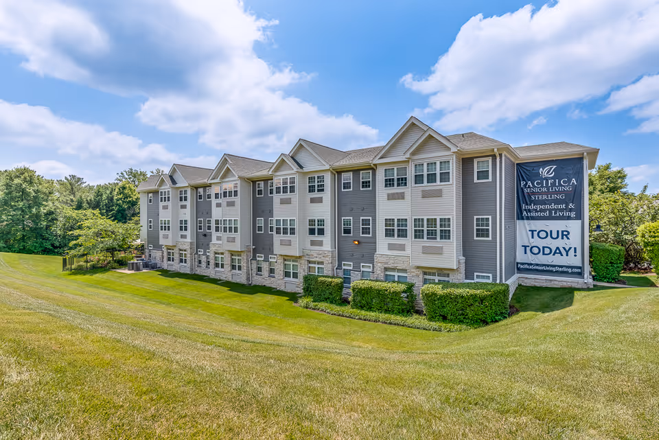 Exterior view of a multi-story senior living building with a large 'Tour Today' banner on the side and a wide grassy lawn in front.