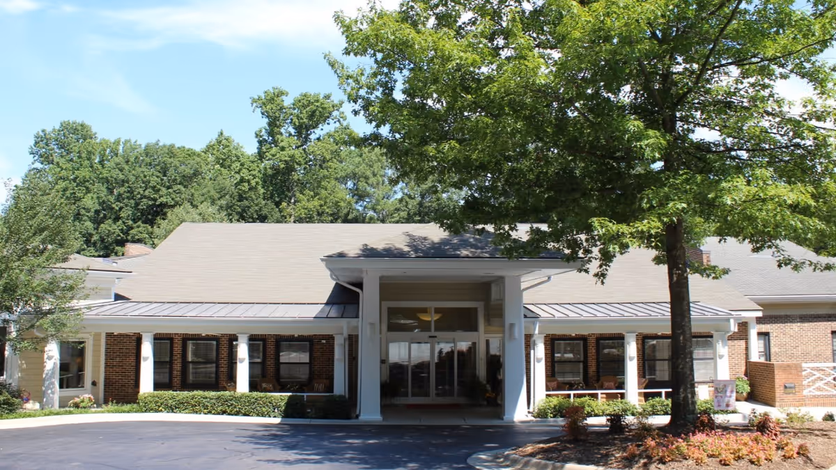 Front exterior view of a single-story brick building with a covered entrance supported by white columns. There are large windows along the front, a tree with green leaves on the right side, and a paved driveway in front. The sky is clear and blue.