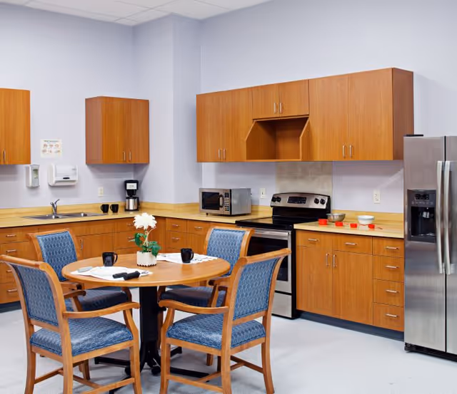A clean and well-lit kitchen area with wooden cabinets, a stainless steel refrigerator, an electric stove with oven, a microwave, a coffee maker, and a round wooden table with four blue cushioned chairs. The table has a small potted plant and some cups on it.