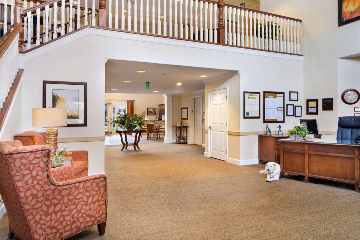 Spacious senior living reception lobby with upholstered chairs, a wooden reception desk, a staircase balcony, and a small dog lying by the desk.