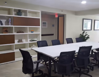 A modern conference room with a long white table surrounded by black office chairs. There is a white shelving unit with decorative items on the left wall and framed pictures on the right wall. A potted plant is placed near the framed pictures.