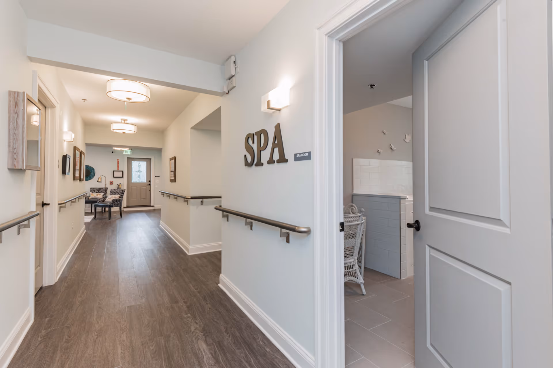 A clean, well-lit hallway with wood flooring and handrails on both sides leading to a seating area with two patterned chairs and a small table near a door with a decorative glass panel. On the right side of the hallway, there is an open door labeled 'SPA' revealing a room with white tiled walls and wicker furniture.