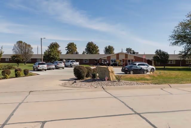 Front exterior view of Falls City Nursing and Rehabilitation Center, a single-story building with a parking lot in front, several cars parked, and a landscaped area with bushes and a large rock sign displaying the facility's name.