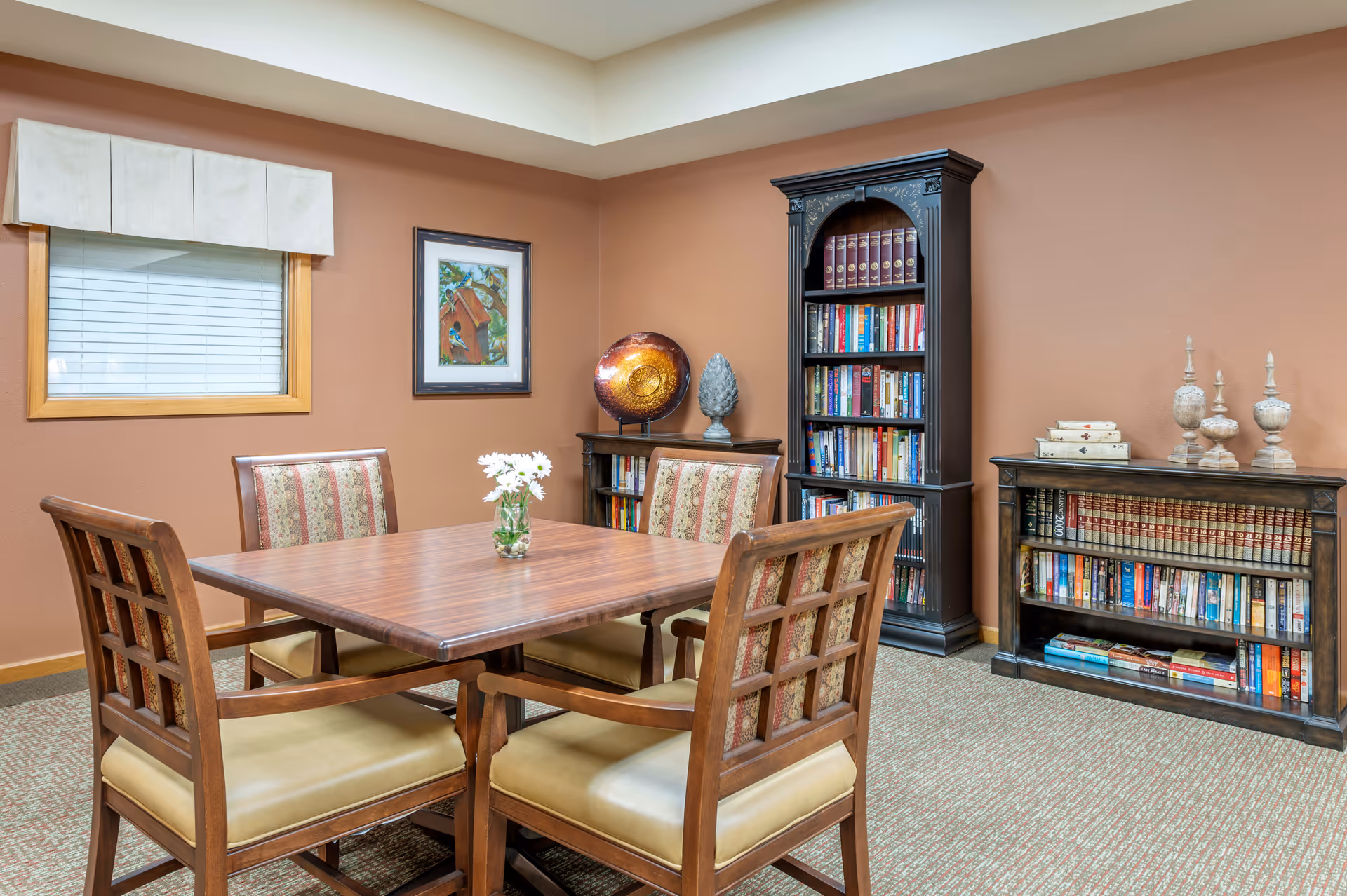 A cozy room with a wooden table surrounded by four cushioned chairs. The walls are painted brown and decorated with a framed picture of a birdhouse. There are two bookshelves filled with books and decorative items, including a large decorative plate and ornamental sculptures. A window with closed blinds and a white valance is on the left wall.