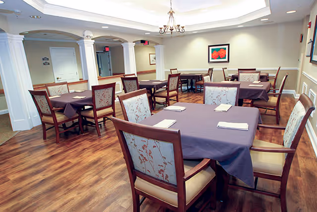 A dining room with several tables covered in purple tablecloths and set with folded white napkins. The room has wooden floors, upholstered chairs with floral patterns, and soft lighting from a chandelier and recessed ceiling lights. There are white columns and beige walls with framed artwork.