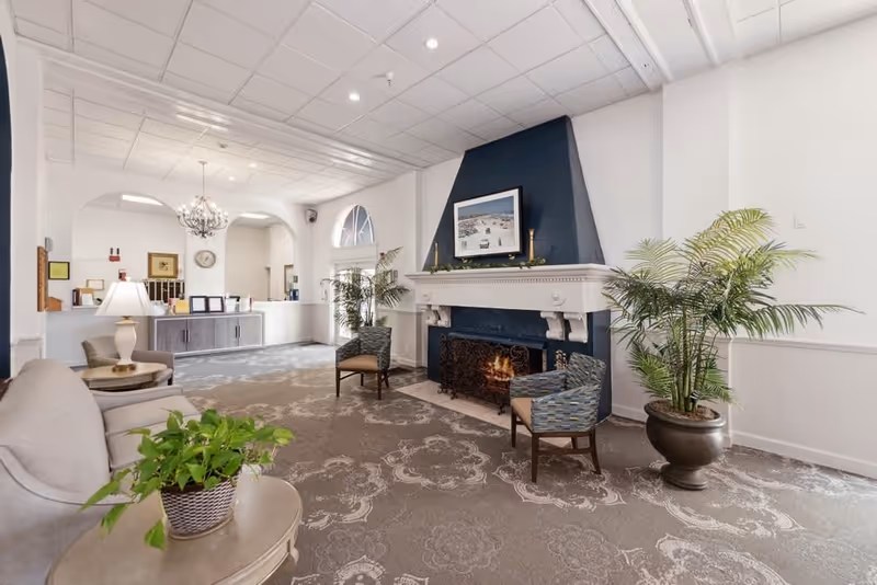 A bright and spacious senior living facility common area featuring a lit fireplace with a dark blue accent wall and white mantel. Two patterned armchairs are placed near the fireplace, along with a large potted plant. A beige sofa and a round side table with a potted plant and lamp are in the foreground. The room has a patterned carpet, white walls, and a chandelier hanging from the ceiling. In the background, there is a reception desk area with framed pictures and a clock on the wall.