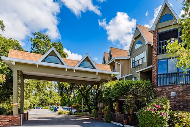 Exterior view of Brookdale Sarasota Midtown senior living facility showing a covered entrance with a peaked roof, surrounded by trees and greenery under a blue sky with scattered clouds.