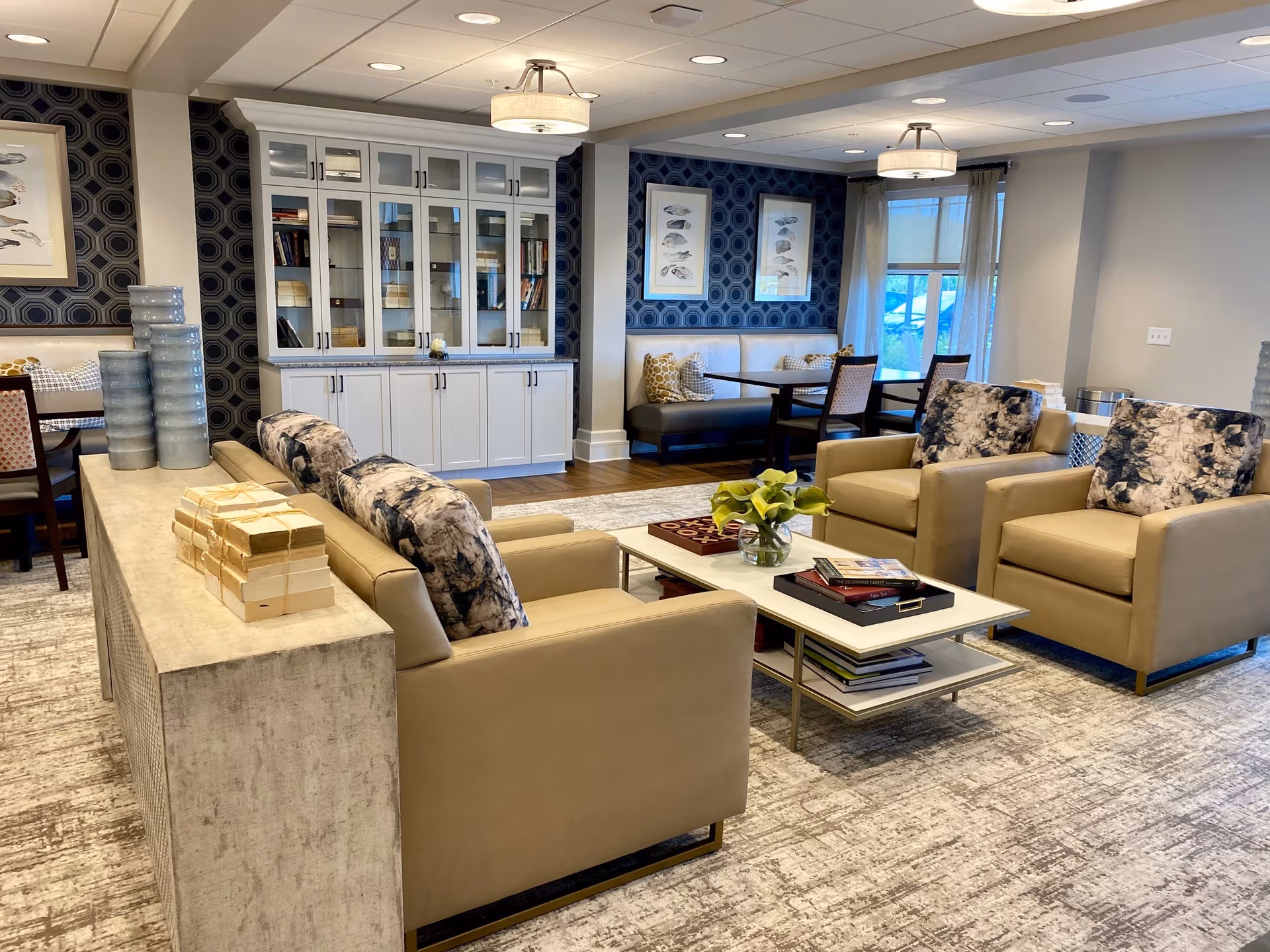 A cozy and well-decorated living room area in a senior living facility featuring beige armchairs with patterned cushions, a beige sofa, a white coffee table with books and a small vase of flowers, and a built-in cabinet with glass doors. The room has patterned wallpaper, framed artwork, and a dining nook with a bench and chairs near a window with sheer curtains.