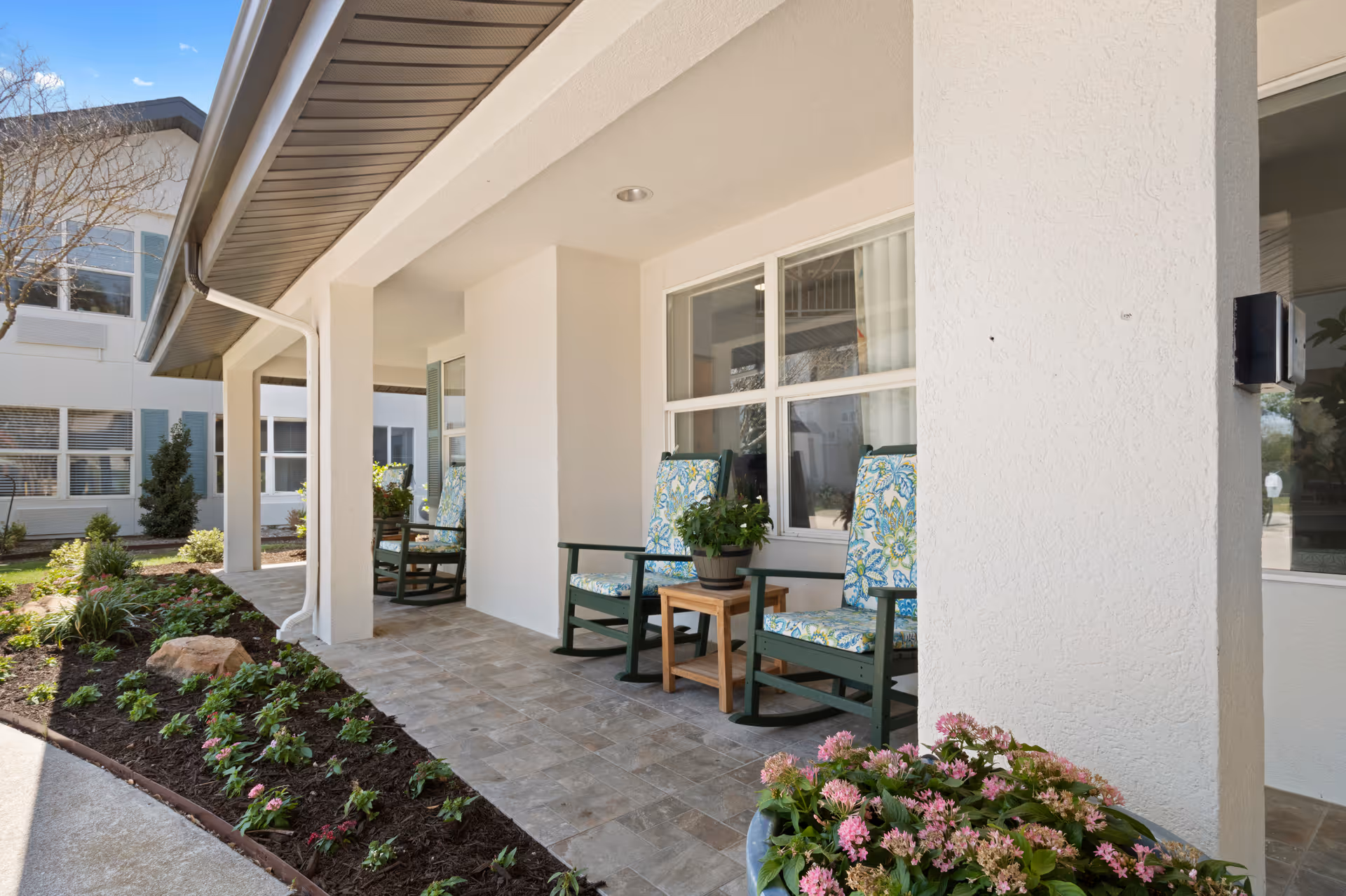 Covered outdoor patio area with three cushioned rocking chairs and a small wooden table with a potted plant. The patio is adjacent to a garden bed with various plants and flowers, and the building exterior is white with large windows.