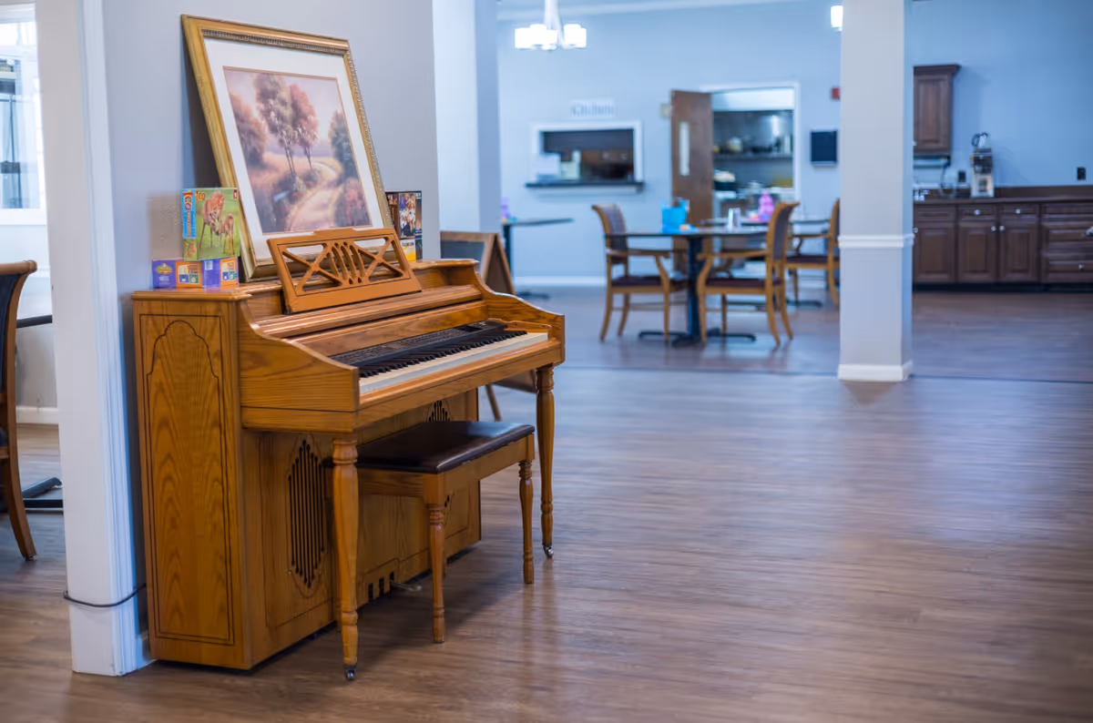 Wooden upright piano with a bench in a spacious senior living common area with dining tables and a kitchen visible in the background.