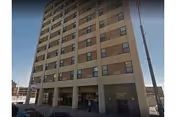 Exterior view of a multi-story apartment building with multiple windows and a covered entrance area at the ground level. The building is situated in an urban environment with a street and parked cars visible in front.
