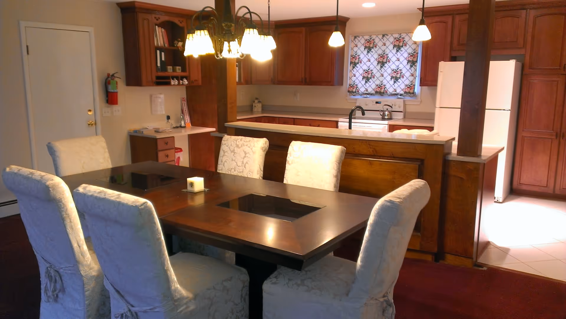 A dining area with a dark wooden table surrounded by six cushioned chairs covered in white fabric. Behind the dining table is a kitchen with wooden cabinets, a white refrigerator, a stove, and a sink. A floral-patterned window curtain is visible above the sink. The room is warmly lit by hanging pendant lights and a chandelier above the dining table.